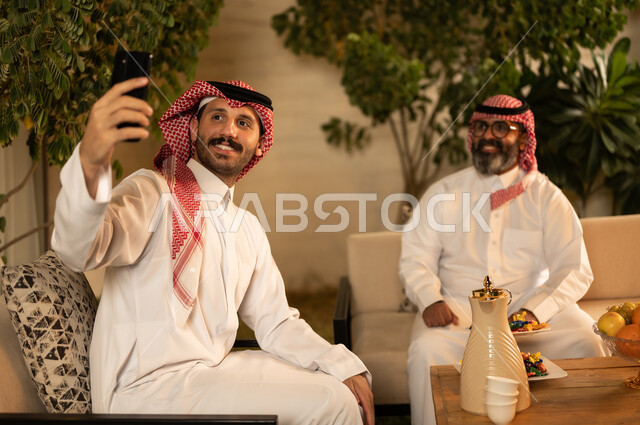 التقاط صورة تذكارية، توثيق اللحظات السعيدة عبر الجوال، رجل عربي خليجي سعودي يرتدي الثوب و الشماغ يجلس برفقة والده يقوم باستخدام الهاتف المحمول، قضاء أوقات عائلية ممتعة ،ايماءات السعادة و السرور