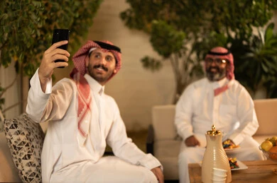 Documenting happy moments through the mobile phone, an Arab Gulf Saudi man wearing a thobe and ghutrah sitting with his father using a mobile phone, taking a commemorative photo, enjoying family time, gestures of happiness and joy.