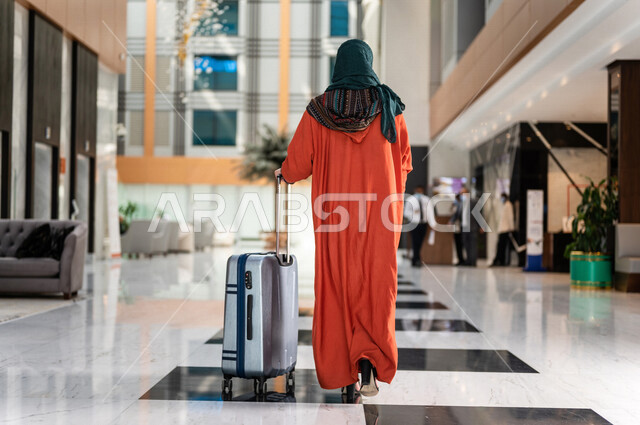 صورة بزاوية من الخلف لسائحة عربية خليجية سعودية تجر حقيبة السفر وتمشي في بهو الفندق ، سيدة أعمال خليجية سعودية ترتدي العباءة الخليجية ، سياحة وفندقة ، قضاء أوقات ممتعة ، رحلة عمل ، سياحة وسفر