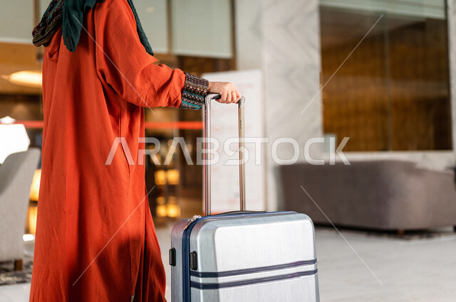 صورة مقربة بزاوية من الخلف لسائحة عربية خليجية سعودية تجر حقيبة السفر وتمشي في بهو الفندق ، سيدة أعمال خليجية سعودية ترتدي العباءة الخليجية ، سياحة وفندقة ، قضاء أوقات ممتعة ، رحلة عمل ، سياحة وسفر