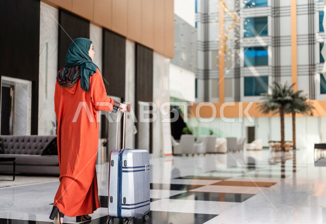 صورة بزاوية من الخلف لسائحة عربية خليجية سعودية تجر حقيبة السفر وتمشي في بهو الفندق ، سيدة أعمال خليجية سعودية ترتدي العباءة الخليجية ، سياحة وفندقة ، قضاء أوقات ممتعة ، رحلة عمل ، سياحة وسفر