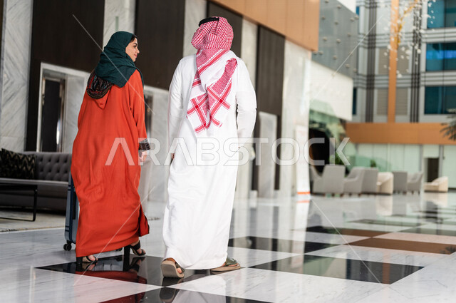 زاوية من الخلف لزوجان عربيان سعوديان سائحان يمشيان ويتجولان في بهو الفندق السياحي ، تمسك الزوجة حقيبة السفر، سياحة وفندقة ، قضاء أجمل الأوقات في أفضل الفنادق الخليجية ، رحلة زوجية ممتعة ، سياحة وسفر، أجواء عائلية مرحة ، عطلة نهاية الأسبوع