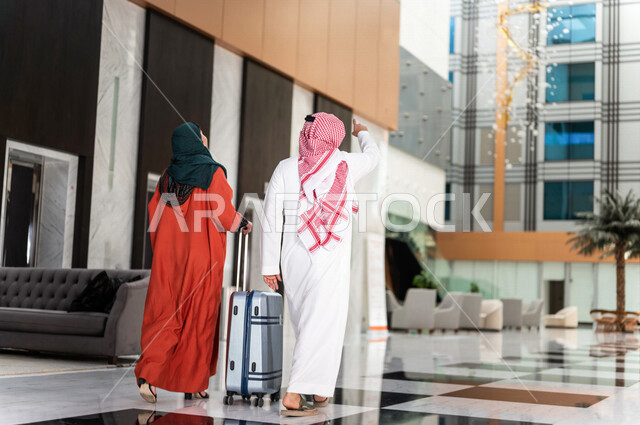 زاوية من الخلف لزوجان عربيان سعوديان سائحان يمشيان ويتجولان في بهو الفندق السياحي ، تمسك الزوجة حقيبة السفر، سياحة وفندقة ، قضاء أجمل الأوقات في أفضل الفنادق الخليجية ، رحلة زوجية ممتعة ، سياحة وسفر، أجواء عائلية مرحة ، عطلة نهاية الأسبوع