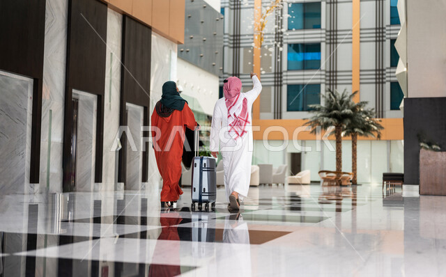 زاوية من الخلف لزوجان عربيان سعوديان سائحان يمشيان ويتجولان في بهو الفندق السياحي ، تمسك الزوجة حقيبة السفر، سياحة وفندقة ، قضاء أجمل الأوقات في أفضل الفنادق الخليجية ، رحلة زوجية ممتعة ، سياحة وسفر، أجواء عائلية مرحة ، عطلة نهاية الأسبوع