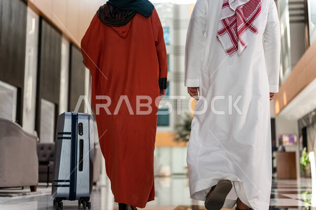زاوية من الخلف لزوجان عربيان سعوديان سائحان يمشيان ويتجولان في بهو الفندق السياحي ، تمسك الزوجة حقيبة السفر، سياحة وفندقة ، قضاء أجمل الأوقات في أفضل الفنادق الخليجية ، رحلة زوجية ممتعة ، سياحة وسفر، أجواء عائلية مرحة ، عطلة نهاية الأسبوع