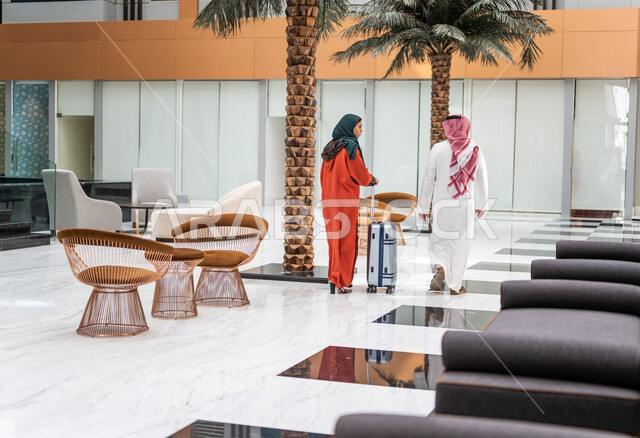 زاوية من الخلف لزوجان عربيان سعوديان سائحان يمشيان ويتجولان في بهو الفندق السياحي ، تمسك الزوجة حقيبة السفر، سياحة وفندقة ، قضاء أجمل الأوقات في أفضل الفنادق الخليجية ، رحلة زوجية ممتعة ، سياحة وسفر، أجواء عائلية مرحة ، عطلة نهاية الأسبوع