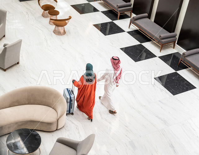 زاوية من الخلف لزوجان عربيان سعوديان سائحان يمشيان ويتجولان في بهو الفندق السياحي ، تمسك الزوجة حقيبة السفر، سياحة وفندقة ، قضاء أجمل الأوقات في أفضل الفنادق الخليجية ، رحلة زوجية ممتعة ، سياحة وسفر، أجواء عائلية مرحة ، عطلة نهاية الأسبوع