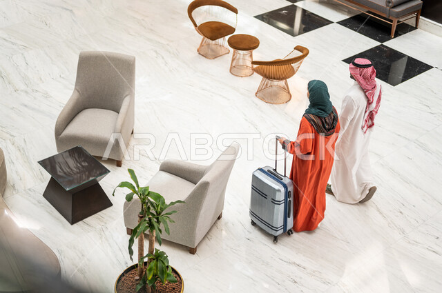 زاوية من الخلف لزوجان عربيان سعوديان سائحان يمشيان ويتجولان في بهو الفندق السياحي ، تمسك الزوجة حقيبة السفر، سياحة وفندقة ، قضاء أجمل الأوقات في أفضل الفنادق الخليجية ، رحلة زوجية ممتعة ، سياحة وسفر، أجواء عائلية مرحة ، عطلة نهاية الأسبوع