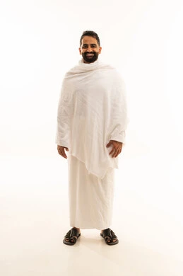 Preparing to perform the rituals of Hajj, standing upright and looking at the camera, a portrait of a smiling Arab Gulf Saudi man wearing the Ihram garment visiting the Sacred House of God in Mecca, full-body, white background.