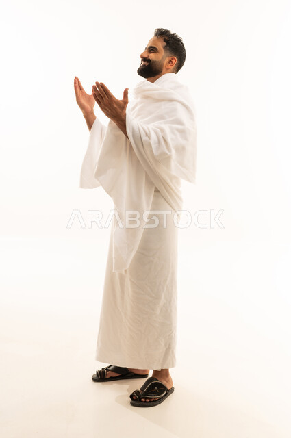 Worship and drawing closer to God, the concept of the pillars of Islam, supplication and pleading to the Lord of the servants, performing the rituals of Hajj and Umrah, a portrait of a Saudi Arabian Gulf man wearing the Ihram clothing raising his hands in gestures of prayer, full-body, white background.