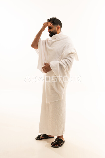 The occurrence of migraine attacks, gestures of headache and feeling unwell, performing the rituals of Hajj and Umrah, the need for treatment and seeking help, a portrait of a smiling Saudi Gulf Muslim man wearing Ihram clothes holding his head with his hands due to severe pain, feeling tired and exhausted, full-body image, white background.