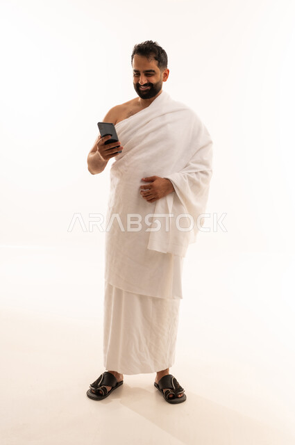 الدعاء عبر الهاتف المحمول, بورتريه لرجل مسلم سعودي خليجي عربي يرتدي ملابس الاحرام يمسك بيدية الجوال، الدليل الالكتروني للحاج و المعتمر, استخدام تطبيقات الجوال للتواصل مع العائلة والأصدقاء, الجسم بالطول الكامل، خلفية بيضاء
