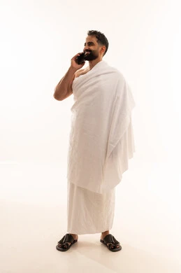 Communicating with family and friends, heading and setting off to visit the Sacred House of God, a portrait of an Arab Gulf Saudi man wearing Ihram clothes holding a mobile phone in his hands making a phone call, performing the pilgrimage and Umrah, using modern technology, a full-body image, white background.
