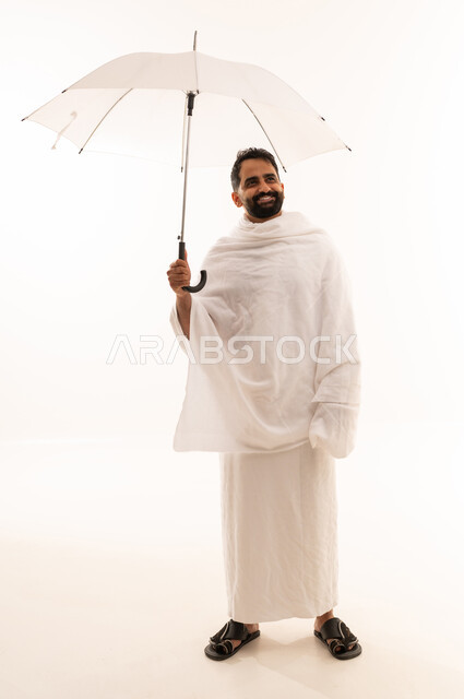 الاستعداد لاداء مناسك الحج والعمره, بورتريه لشاب خليجي عربي سعودي يرتدي ثياب الإحرام يمسك بيده المظله الشمسيه لمقاومه المناخ الحار، صوره الجسم بالطول الكامل، خلفية بيضاء