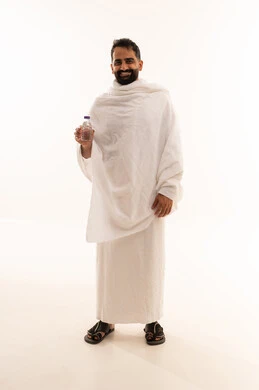 Looking at the camera with expressions of joy, the fifth pillar of Islam for those who are able, standing and drinking Zamzam water after performing the obligatory and voluntary prayers in the holy city of Mecca, preparing to perform the rituals of Hajj and Umrah and getting closer to God through good deeds, a portrait of an Arab Gulf Saudi man wearing the Ihram garment, full-body, white background.