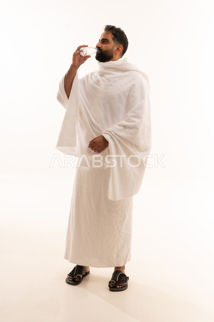 Performing the rituals of Hajj and Umrah, increasing fluid intake to keep the body hydrated, a portrait of an Arab Saudi Gulf man wearing the Ihram attire drinking fresh water, quenching his thirst with Zamzam water, full-body shot, white background.