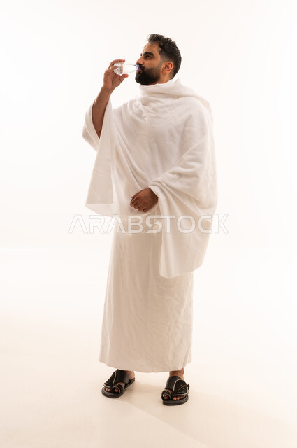 Increasing fluid intake to keep the body hydrated, performing the rituals of Hajj and Umrah, a portrait of an Arab Saudi Gulf man wearing the Ihram attire drinking fresh water, quenching his thirst with Zamzam water, full-body shot, white background.
