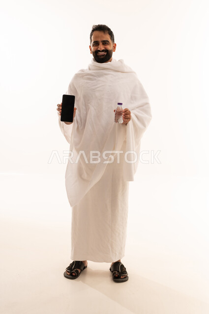 استخدام الأجهزة التقنية الحديثه في التواصل، عرض تطبيقات وبرامج دليل الحاج والمعتمر، بورتريه لشاب خليجي عربي سعودي يرتدي ثياب الإحرام يمسك الهاتف المحمول و زجاجة مياه، صوره الجسم بالطول الكامل، خلفية بيضاء