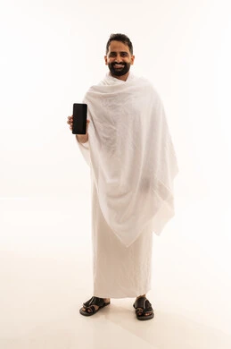عرض تطبيقات وبرامج دليل الحاج والمعتمر، بورتريه لشاب خليجي عربي سعودي يرتدي ثياب الإحرام يمسك الهاتف المحمول، استخدام الأجهزة التقنية الحديثه في التواصل، صوره الجسم بالطول الكامل، خلفية بيضاء