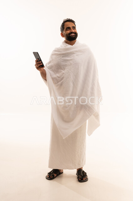 الدليل الالكتروني للحاج و المعتمر، النظر الى شيء ما، الدعاء عبر الهاتف المحمول, بورتريه لرجل مسلم سعودي خليجي عربي يرتدي ملابس الاحرام ويجري مكالمة فيديو, استخدام تطبيقات الجوال للتواصل مع العائلة والأصدقاء, الجسم بالطول الكامل، خلفية بيضاء
