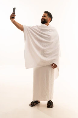توثيق اللحظات السعيدة في مكة المكرمة، استخدام أجهزة تقنية حديثة، أداء مناسك الحج والعمرة، بورتريه لرجل عربي خليجي سعودي يرتدي ملابس الاحرام ويقوم بالتقاط صور سيلفي تذكارية عبر الهاتف المحمول، الجسم بالطول الكامل، خلفية بيضاء