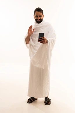 اجراء مكالمة فيديوعبر الهاتف المحمول, رفع كف اليد بتعابير التحية و السلام، بورتريه لرجل مسلم سعودي خليجي عربي يرتدي ملابس الاحرام يمسك بيدية الجوال, الدليل الالكتروني للحاج و المعتمر, استخدام الموبيل الجوال للتواصل مع العائلة والأصدقاء, الجسم بالطول الكامل، خلفية بيضاء