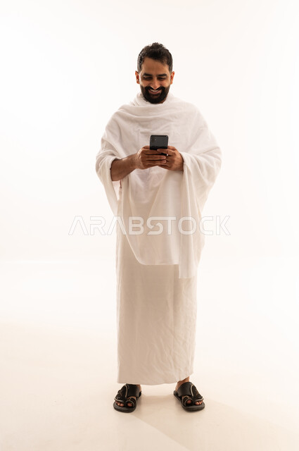 الدعاء عبر الهاتف المحمول, بورتريه لرجل مسلم سعودي خليجي عربي يرتدي ملابس الاحرام ويجري مكالمة فيديو, الدليل الالكتروني للحاج و المعتمر, استخدام تطبيقات الجوال للتواصل مع العائلة والأصدقاء, الجسم بالطول الكامل، خلفية بيضاء