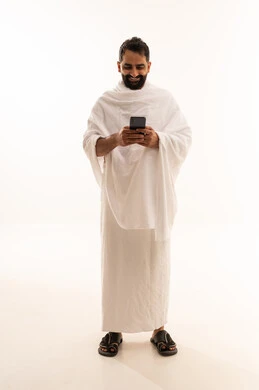 الدعاء عبر الهاتف المحمول, بورتريه لرجل مسلم سعودي خليجي عربي يرتدي ملابس الاحرام ويجري مكالمة فيديو, الدليل الالكتروني للحاج و المعتمر, استخدام تطبيقات الجوال للتواصل مع العائلة والأصدقاء, الجسم بالطول الكامل، خلفية بيضاء