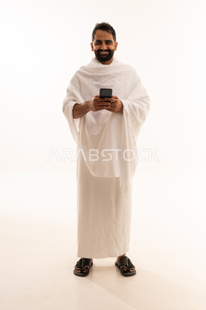 النظر الى الكاميرا بتعابير السرور، الدعاء عبر الهاتف المحمول, بورتريه لرجل مسلم سعودي خليجي عربي يرتدي ملابس الاحرام ويجري مكالمة فيديو, الدليل الالكتروني للحاج و المعتمر, استخدام تطبيقات الجوال للتواصل مع العائلة والأصدقاء, الجسم بالطول الكامل، خلفية بيضاء