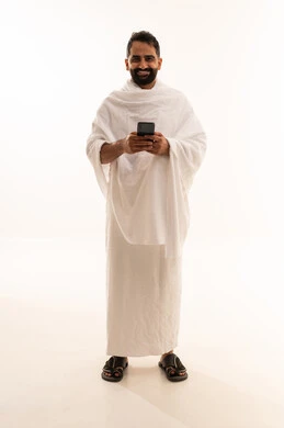 Looking at the camera with expressions of joy, praying via mobile phone, a portrait of a Saudi Arabian Gulf Arab Muslim man wearing Ihram clothing and making a video call, the electronic guide for pilgrims and Umrah performers, using mobile applications to communicate with family and friends, full-body, white background.