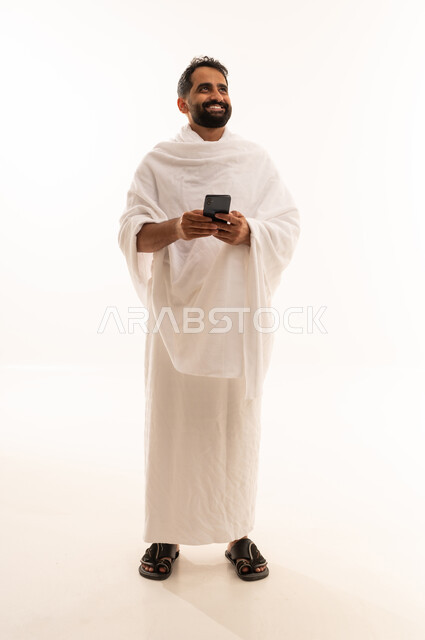 النظر الى شيء ما، الدعاء عبر الهاتف المحمول, بورتريه لرجل مسلم سعودي خليجي عربي يرتدي ملابس الاحرام ويجري مكالمة فيديو, الدليل الالكتروني للحاج و المعتمر, استخدام تطبيقات الجوال للتواصل مع العائلة والأصدقاء, الجسم بالطول الكامل، خلفية بيضاء