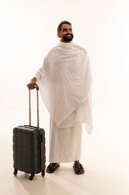 النظر الى شيء ما بتعابير السرور، الاستعداد للتوجه و الانطلاق لزيارة بيت الله الحرام، بورتريه لرجل خليجي عربي سعودي مبتسم يرتدي ملابس الاحرام ويمسك بيده حقيبة السفر، اداء فريضة الحج والعمرة، الجسم بالطول الكامل، خلفية بيضاء