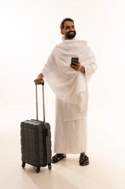 Looking at something, using mobile applications to communicate with family and friends, preparing to head out and set off to visit the House of God, a portrait of a Gulf Arab man wearing Ihram clothing holding a mobile phone and a travel bag, full-body, white background.