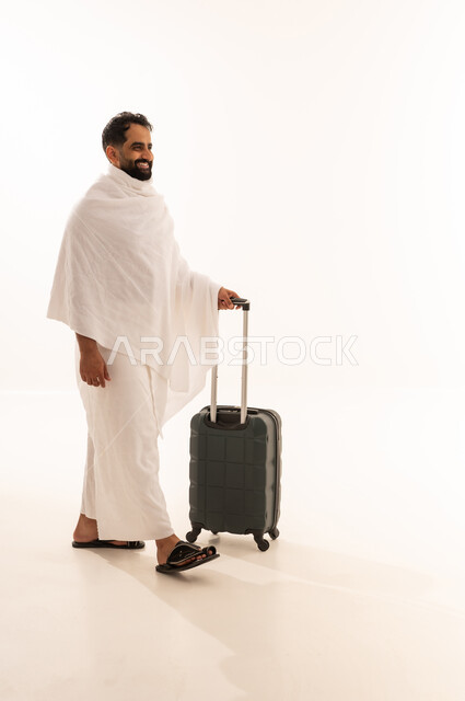 التقرب الى الله من خلال أداء فريضة الحج والعمرة، السفر والترحال لزيارة بيت الله الحرام، بورتريه من الجانب لرجل مسلم خليجي عربي سعودي مبتسم يرتدي ملابس الاحرام يمسك بيده حقيبة سفر، النظر بإيماءات السرور والسعادة، صورة الجسم بالكامل، خلفية بيضاء