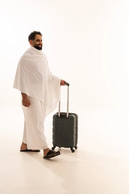 التقرب الى الله من خلال أداء فريضة الحج والعمرة، السفر والترحال لزيارة بيت الله الحرام، بورتريه من الجانب لرجل مسلم خليجي عربي سعودي مبتسم يرتدي ملابس الاحرام يمسك بيده حقيبة سفر، النظر بإيماءات السرور والسعادة، صورة الجسم بالكامل، خلفية بيضاء