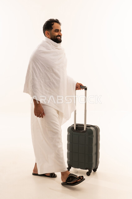 السفر والترحال لزيارة بيت الله الحرام، بورتريه من الجانب لرجل مسلم خليجي عربي سعودي مبتسم يرتدي ملابس الاحرام يمسك بيده حقيبة سفر ، التقرب الى الله من خلال أداء فريضة الحج والعمرة، النظر بإيماءات السرور والسعادة، صورة الجسم بالكامل، خلفية بيضاء