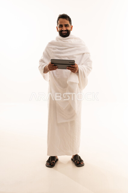 النظر الى الكاميرا بتعابير السرور، التواصل مع الاهل والاصدقاء باستخدام التابلت، بورتريه لرجل خليجي عربي سعودي يرتدي ثياب الإحرام يقوم بتصفح الجهاز اللوحي، صوره الجسم بالطول الكامل، خلفية بيضاء