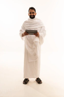 النظر الى الكاميرا بتعابير السرور، التواصل مع الاهل والاصدقاء باستخدام التابلت، بورتريه لرجل خليجي عربي سعودي يرتدي ثياب الإحرام يقوم بتصفح الجهاز اللوحي، صوره الجسم بالطول الكامل، خلفية بيضاء
