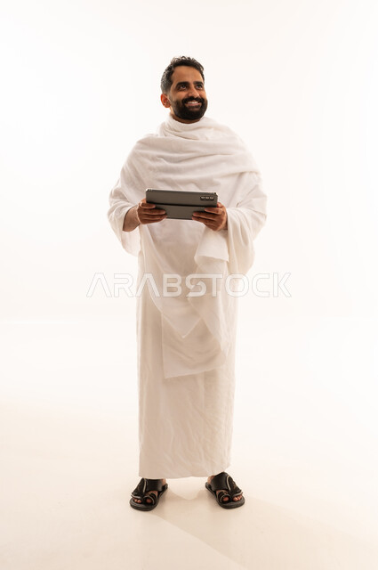 النظر الى شيء ما بتعابير السرور، التواصل مع الاهل والاصدقاء باستخدام التابلت، بورتريه لرجل خليجي عربي سعودي يرتدي ثياب الإحرام يقوم بتصفح الجهاز اللوحي، صوره الجسم بالطول الكامل، خلفية بيضاء