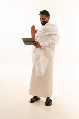 اجراء مكالمة فيديو، رفع كف اليد بتعابير التحية و السلام، التواصل مع الاهل والاصدقاء باستخدام التابلت، بورتريه لرجل خليجي عربي سعودي يرتدي ثياب الإحرام يقوم بتصفح الجهاز اللوحي، صوره الجسم بالطول الكامل، خلفية بيضاء
