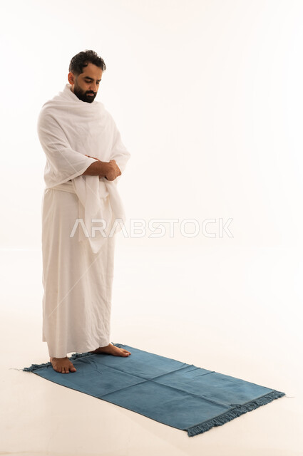 الخشوع والتقرب إلى الله، الركن الثاني من أركان الإسلام، أداء مناسك الحج والعمرة،بورتريه لرجل عربي خليجي يرتدي ملابس الإحرام يقف باستقامة يؤدي فريضة الصلاة، مفهوم العبادة والحفاظ على الصلوات، صورة الجسم بالطول الكامل، خلفية بيضاء