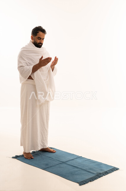 Raising the hands upwards and praying, seeking forgiveness and mercy from God Almighty, worship and obedience, and increasing the remembrance of God, performing the rituals of Hajj and Umrah, a portrait of an Arab Gulf man wearing Ihram clothing praying to God, full-body, white background.