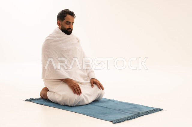 الجلوس للتشهد على السجادة، الخشوع في الصلاة والتضرع إلى الله، بورتريه لرجل عربي سعودي خليجي يرتدي لباس الاحرام يجلس على سجادة الصلاة يؤدي الفريضة، أداء مناسك الحج والعمرة، التقرب إلى الله بالعبادة والطاعة، خلفية بيضاء