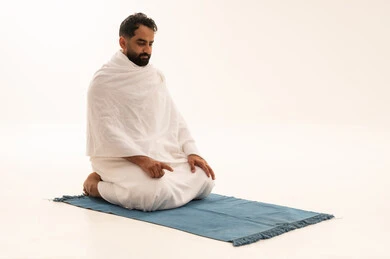 Humility in prayer and supplication to God, sitting for the testimony on the prayer rug, a portrait of an Arab Saudi Gulf man wearing the Ihram garment sitting on the prayer rug performing the obligatory prayer, performing the rituals of Hajj and Umrah, drawing closer to God through worship and obedience, white background.