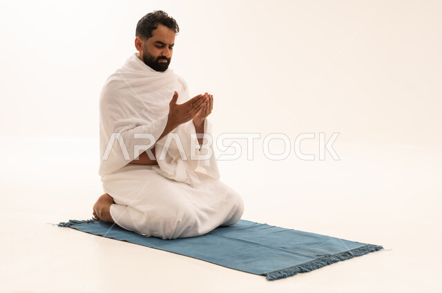 Drawing closer to God through diligence in worship and performing the rituals of Hajj and Umrah, performing the obligatory prayers on time, a portrait of an Arab Saudi Gulf man wearing the Ihram garment sitting on a prayer rug, raising his hands and praying to God, gestures indicating supplication and humility, white background.