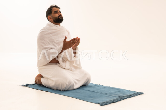Performing the obligatory prayers on time, getting closer to God through diligence in worship and performing the rituals of Hajj and Umrah, a portrait of an Arab Saudi Gulf man wearing the Ihram garment sitting on a prayer rug, raising his hands and praying to God, gestures indicating supplication and humility, white background.
