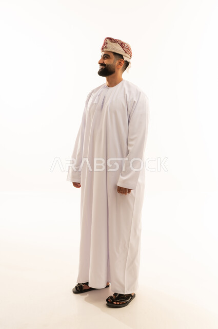 تعابير الثقة بالنفس، النظر الى شيء ما بايماءات السرور، مفهوم الرجولة والوقار، الاهتمام بالمظهر الخارجي، بورتريه لرجل عربي خليجي عماني يرتدي الدشداشة والعمامة يقف باستقامة، صورة الجسم بالطول الكامل، خلفية بيضاء