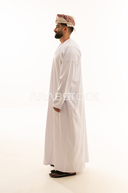 The concept of handsome for men, standing upright and straight, a side portrait of a smiling young Arab Gulf Omani man wearing a dishdasha and turban looking at something with gestures of joy and happiness, attention to elegance and external appearance, full-body image, white background.