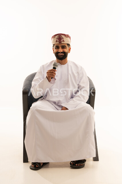 النظر للكاميرا بتعابير السرور، ايماءات وتعابير وجه و يدين تعبر عن الغناء والطرب، فن إلقاء الخطابات والندوات، التدريب الصوتي وتقديم البرامج الاذاعيه، بورتريه لرجل عربي خليجي عماني يرتدي الدشداشة و العمامة يجلس على الكرسى يمسك بيدية الميكروفون، خلفية بيضاء