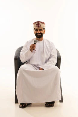 Looking at the camera with expressions of joy, gestures and facial and hand expressions that convey singing and music, the art of delivering speeches and seminars, vocal training and presenting radio programs, a portrait of an Arab Gulf Omani man wearing a dishdasha and a turban sitting on a chair holding a microphone in his hands, with a white background.